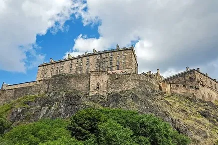 Edinburgh Castle on a rocky hill with green trees below, under a partly cloudy blue sky.
