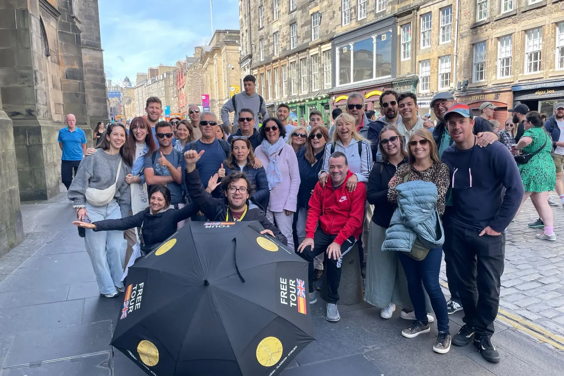 A diverse group of smiling tourists and their guide holding a 'Free Tour' umbrella on a cobblestone street in Edinburgh.
