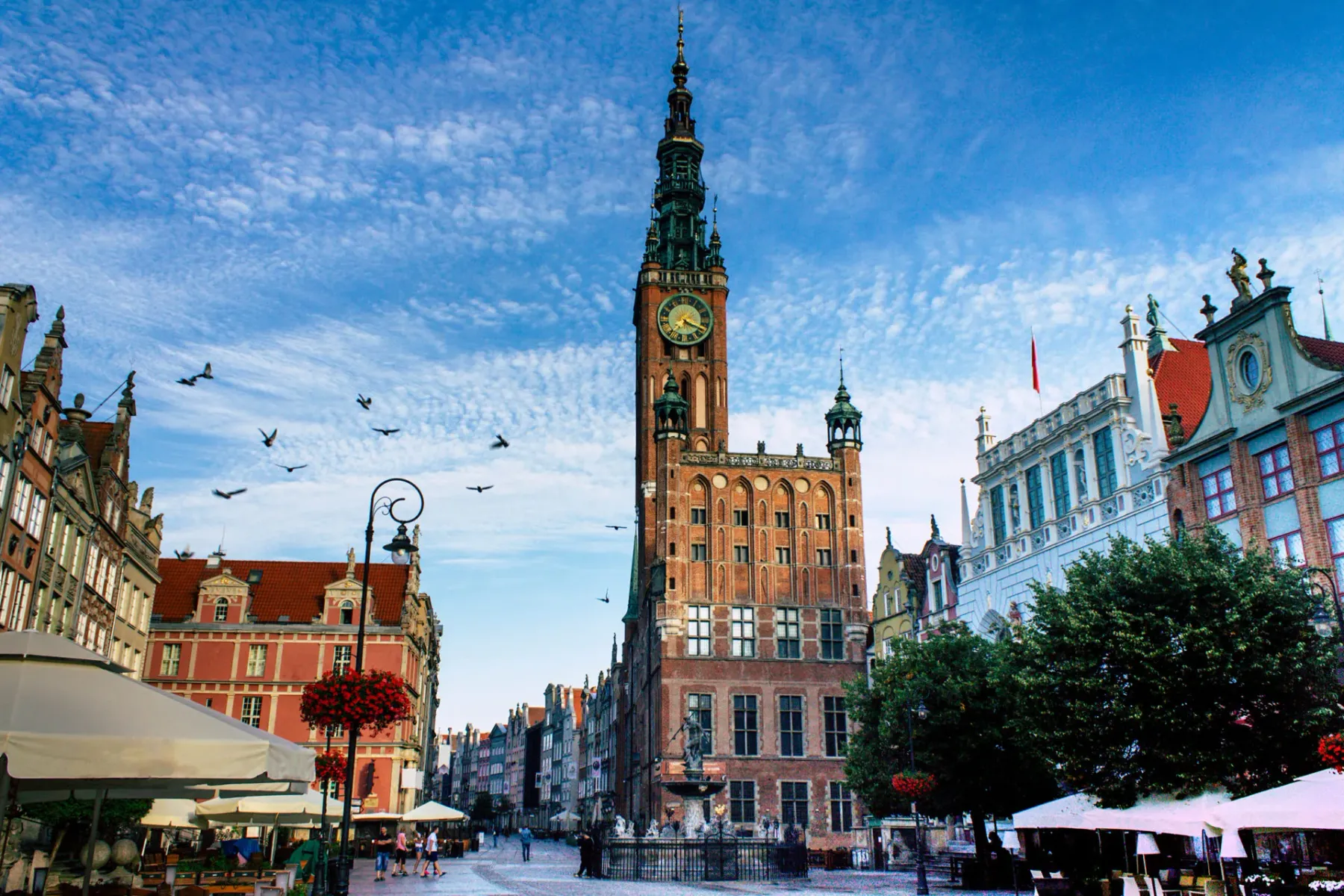 Main Town Hall of Gdańsk with its tall spire and clock, surrounded by historic buildings and people walking on a sunny day.