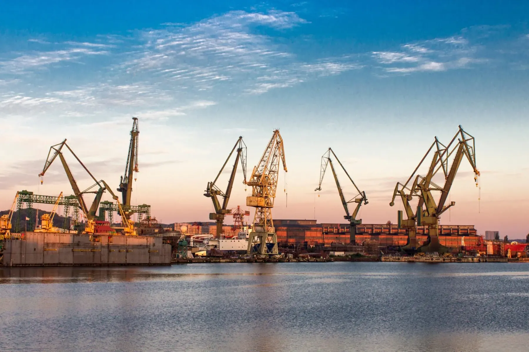 Multiple large industrial cranes and buildings of a shipyard under a partly cloudy sky, with reflections in the water at s...