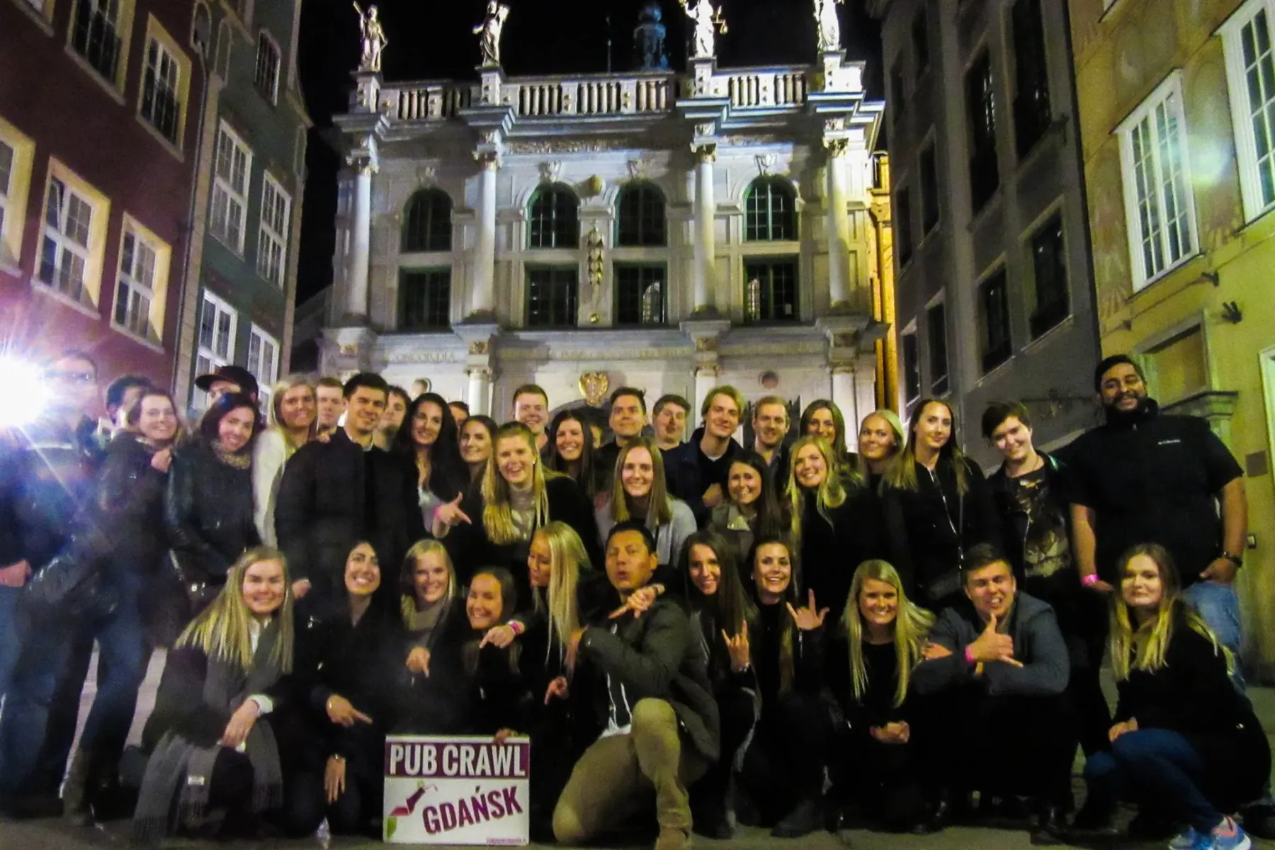 A large group of young adults posing at night in front of an ornate historic building in Gdansk, holding a 'Pub Crawl Gdań...