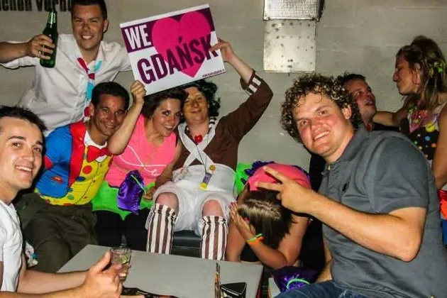 Group of smiling people, some in costumes, holding a 'We <3 Gdańsk' sign in an indoor setting with drinks.