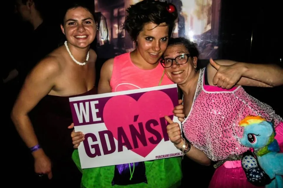 Three smiling women pose with a white sign displaying 'WE ❤️ GDAŃSK' and 'pubcrawlgdansk.pl' against a dark background.