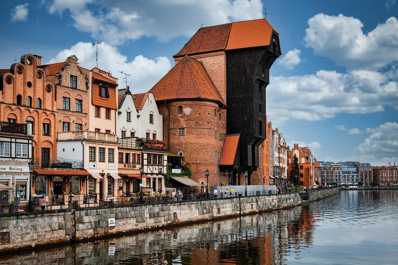 Historic wooden crane building (Żuraw) and traditional brick buildings lining the Motława River under a cloudy sky in Gdansk.