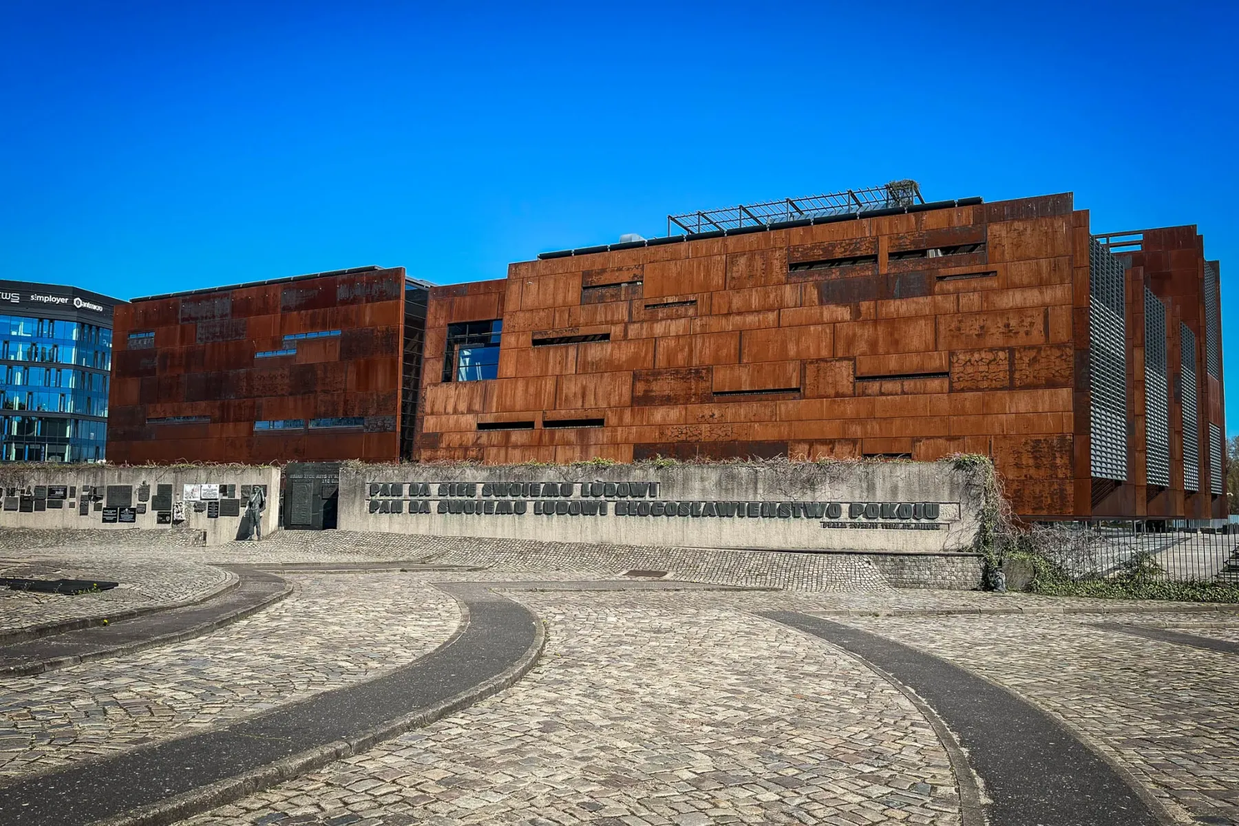 Building with rusted metal facade, cobblestone plaza, and concrete memorial wall with plaques and Polish text. Clear blue ...