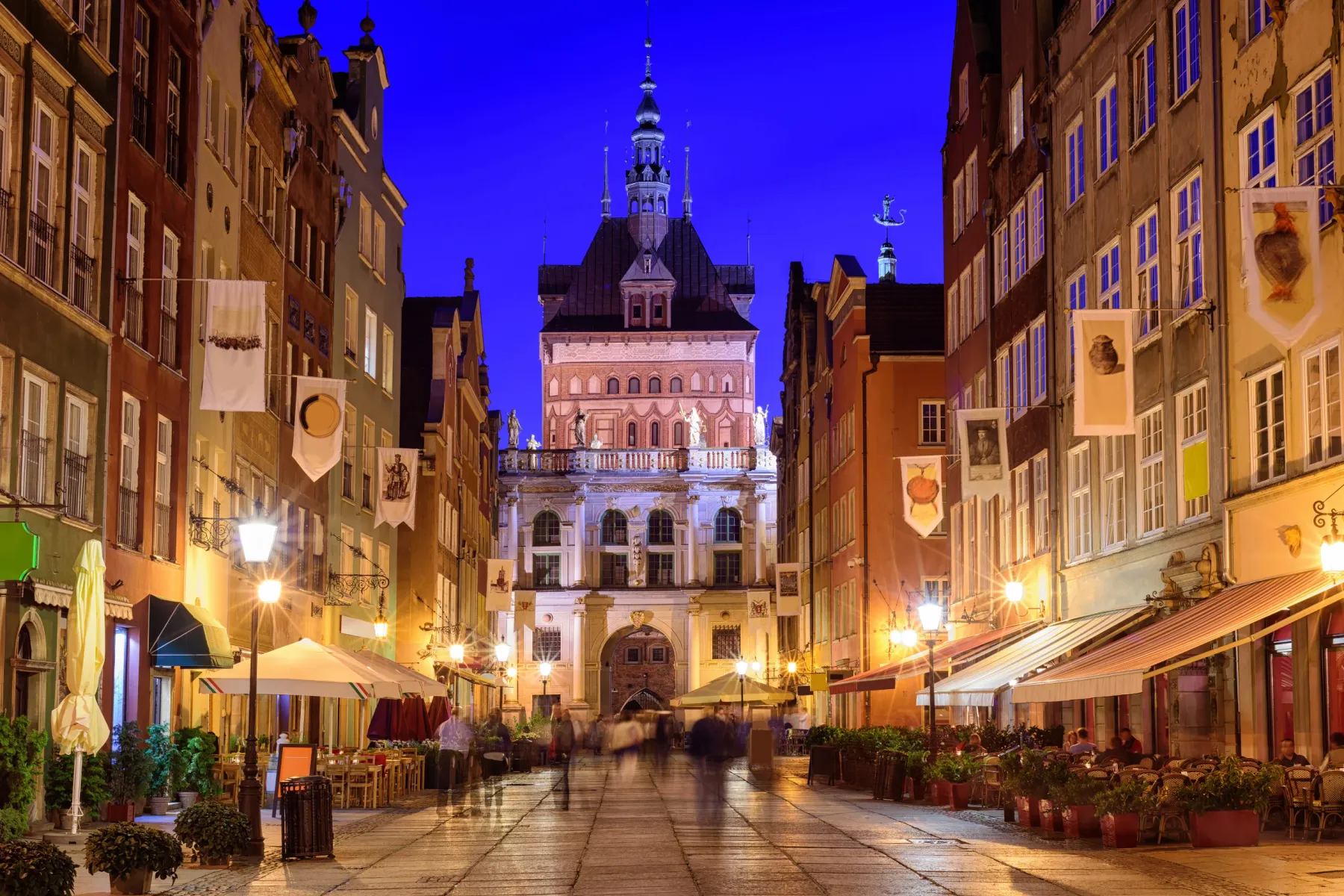 Gdańsk's Długa Street at dusk, lined with historical buildings and outdoor cafes. The illuminated Main Town Hall stands at...
