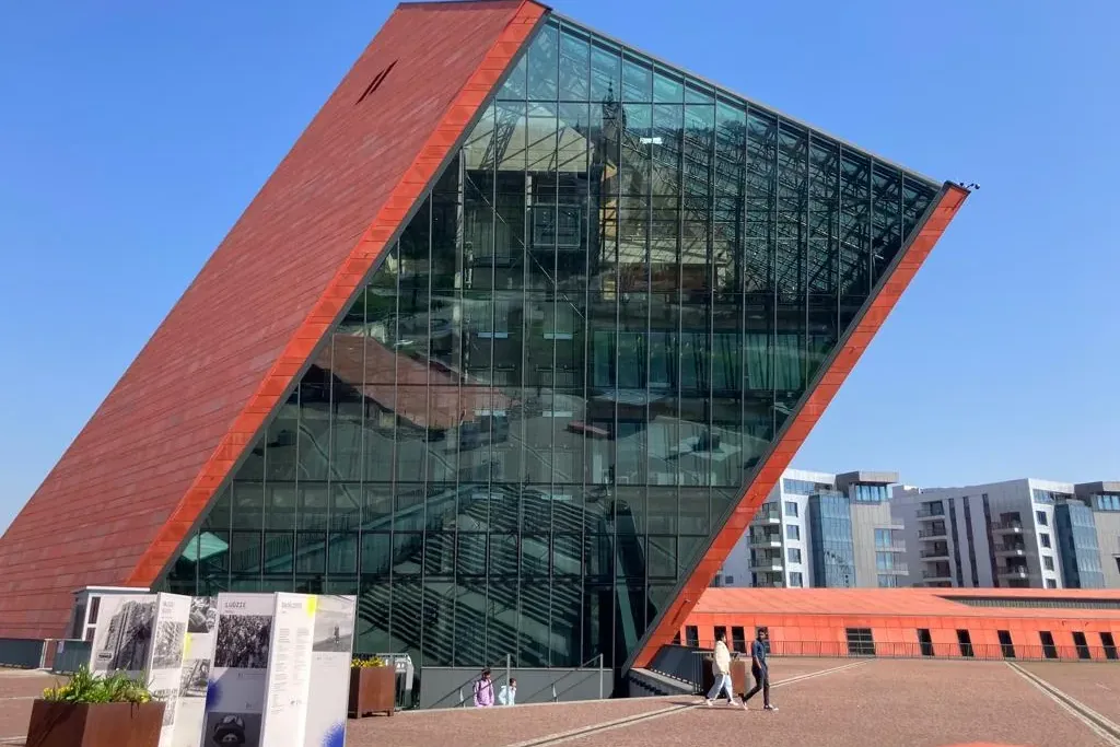 Modern building with a large red-brown angled wall and a reflective glass facade, people walking on a paved plaza.