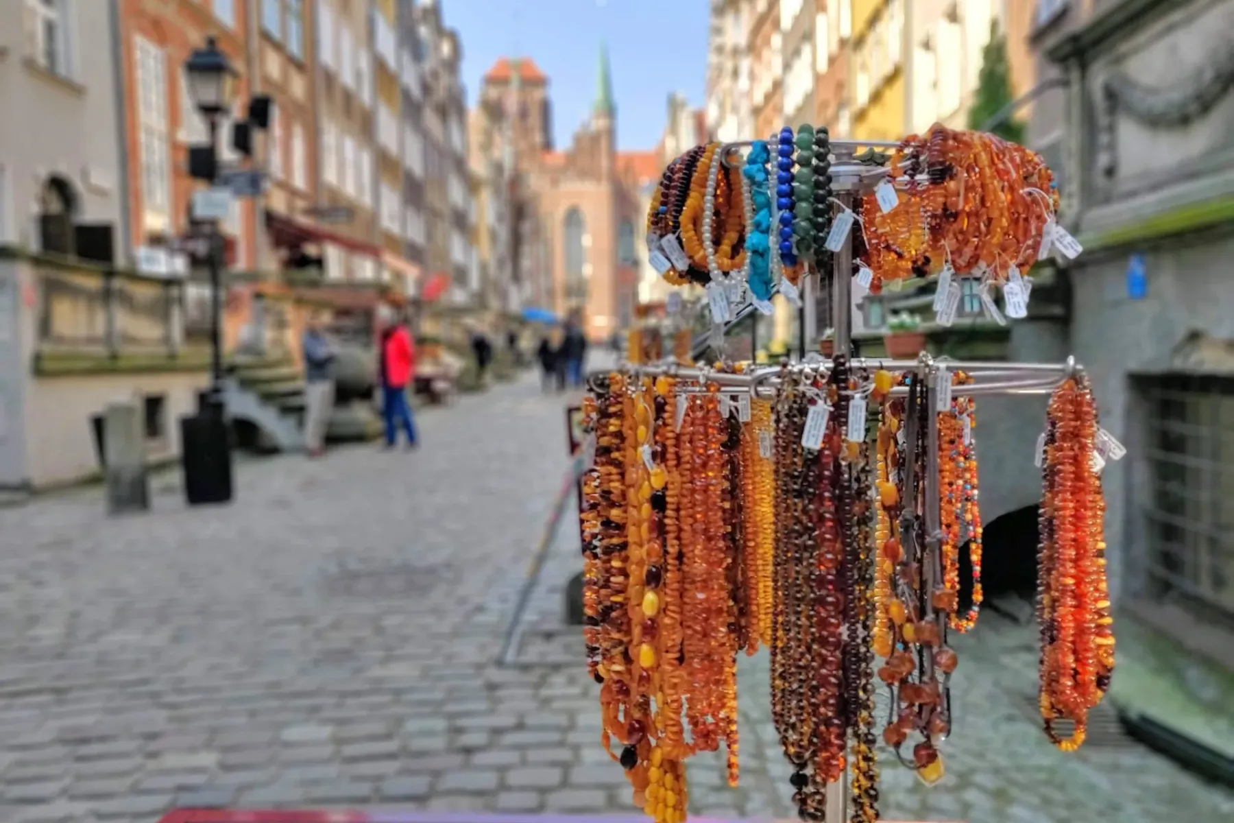 A street stall displaying numerous amber necklaces and bracelets in various shades, with a blurred old town street of Gdań...