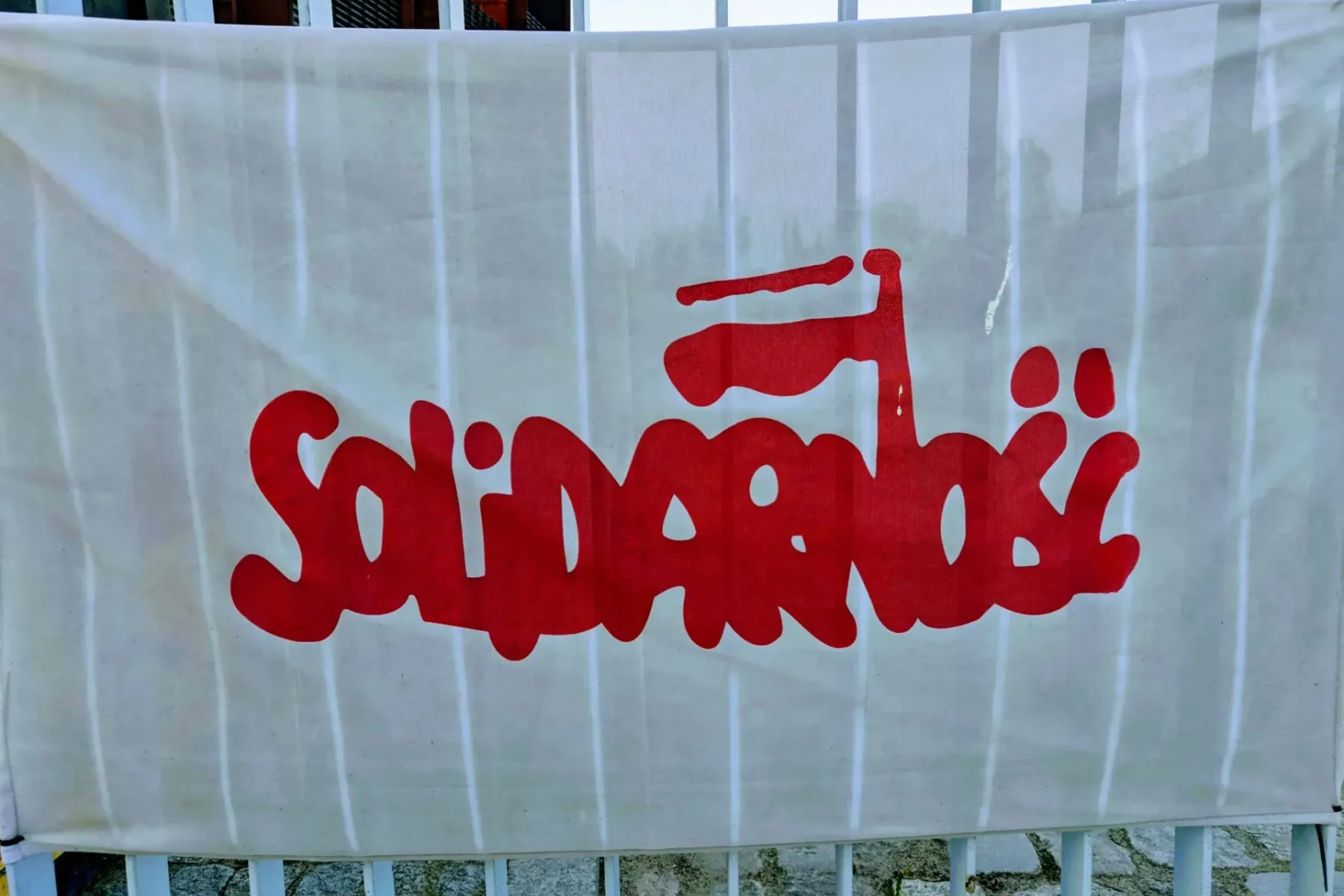 White banner with red Solidarność logo, featuring a stylized Polish flag. The banner is affixed to a white metal fence.