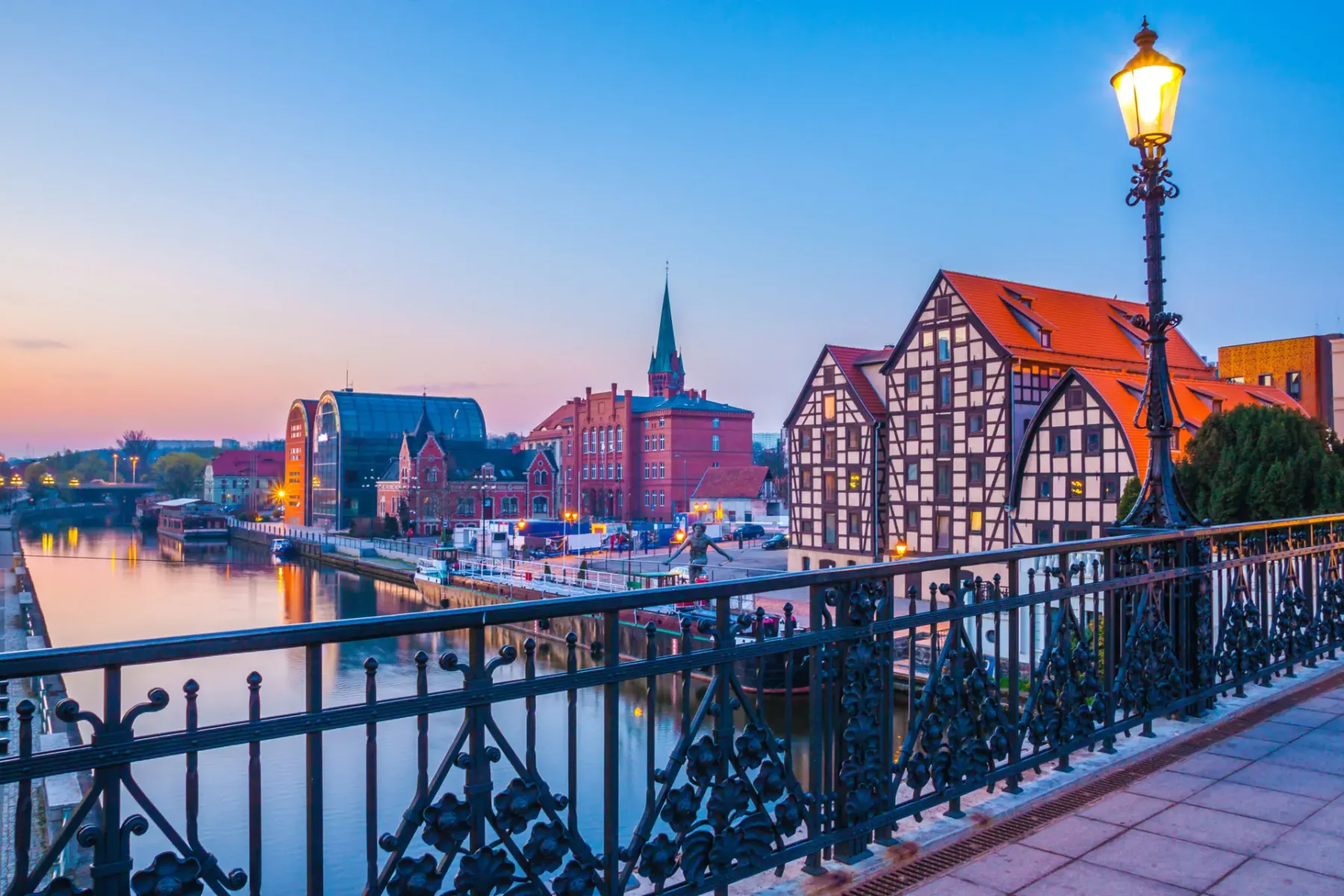 View of historic granaries and other buildings along the Brda River in Bydgoszcz at dusk, with a decorative bridge railing...