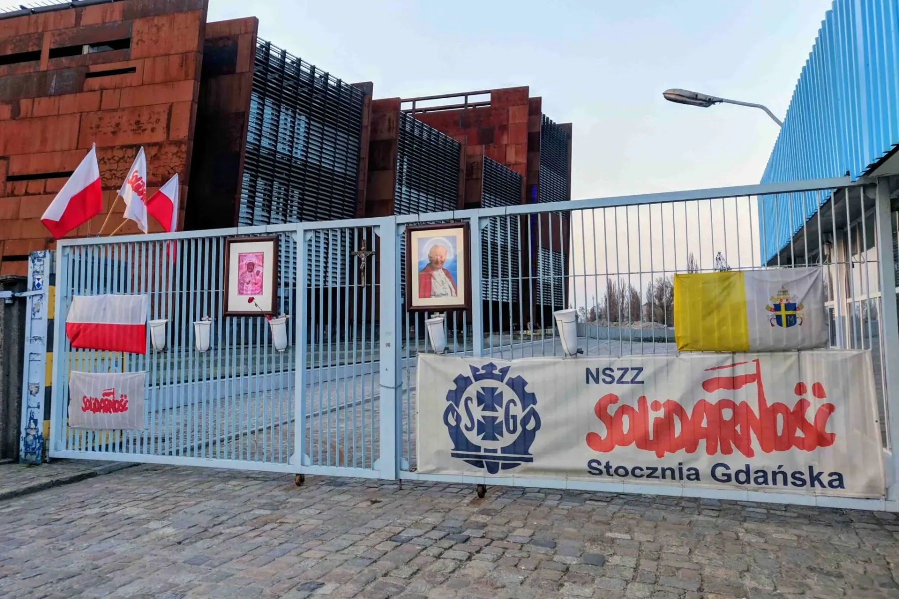 Metal gate with Polish and Vatican flags, Solidarity banners, and framed images in front of a rust-colored building.