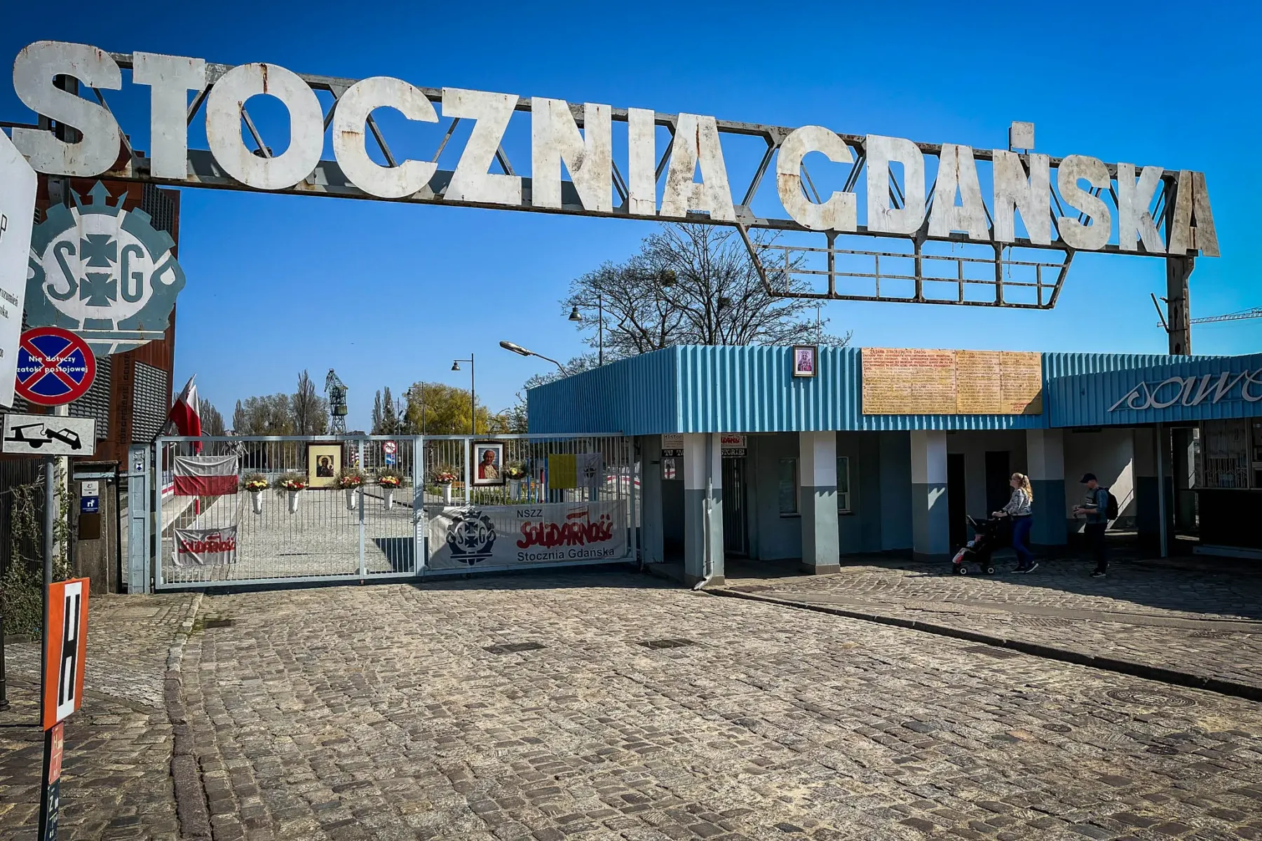 Large metal sign "STOCZNIA GDAŃSKA" over a gated entrance with a Solidarity banner and Polish flags, cobblestone ground.
