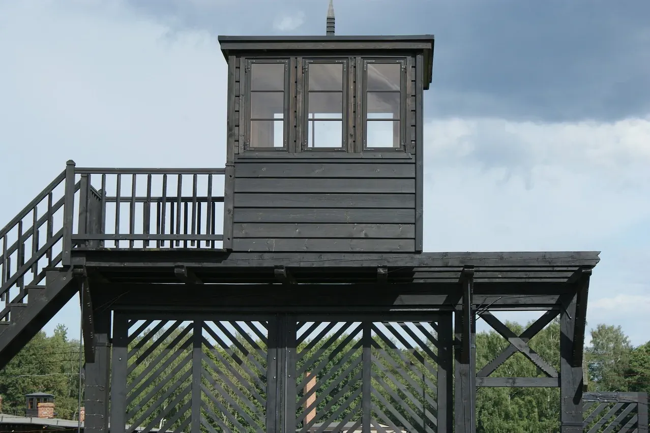 Dark wooden guard tower with a small room and three windows, a staircase, and a lattice fence below, against a cloudy sky.