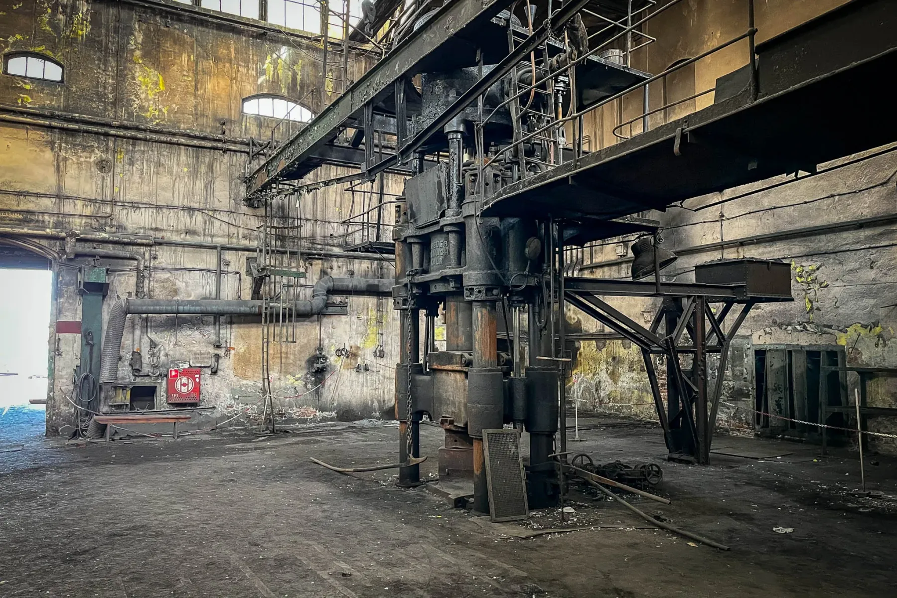 Derelict industrial hall with a large, dark hydraulic press machine in the center, dirty floor, peeling walls, and high wi...