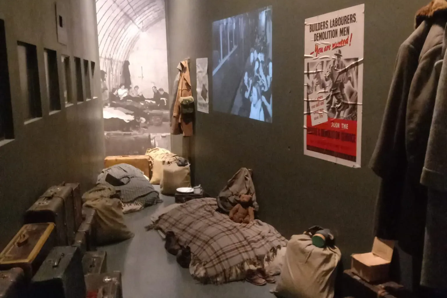 Dark museum exhibit room depicting a wartime shelter with stacked suitcases, rumpled blankets, clothes, and a propaganda p...