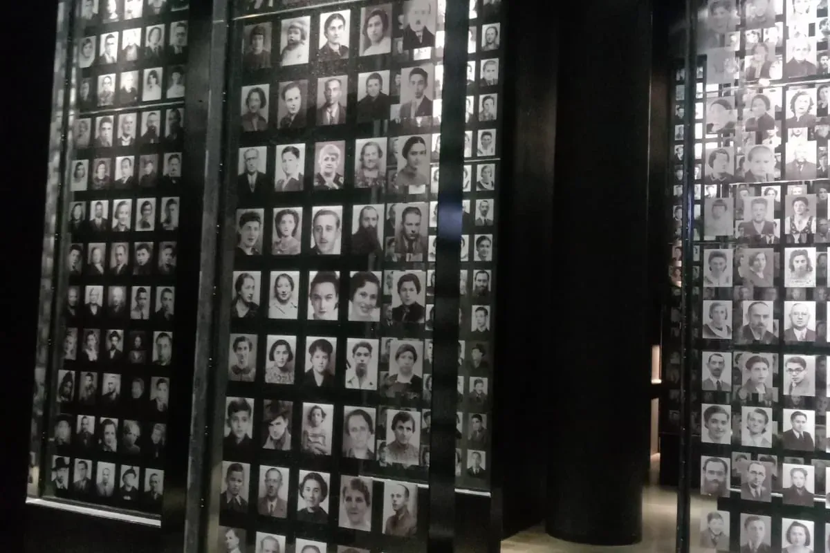 Dark museum hall with tall, illuminated display panels filled with small black and white photographic portraits of individ...