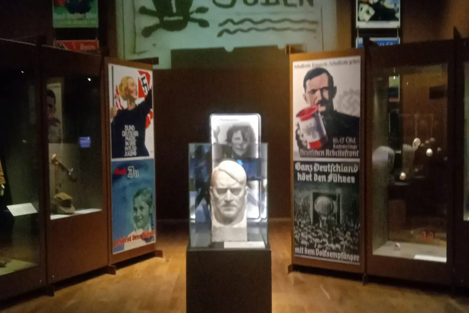 Museum exhibit with a bust on a pedestal, surrounded by glass display cases and historical propaganda posters, including '...