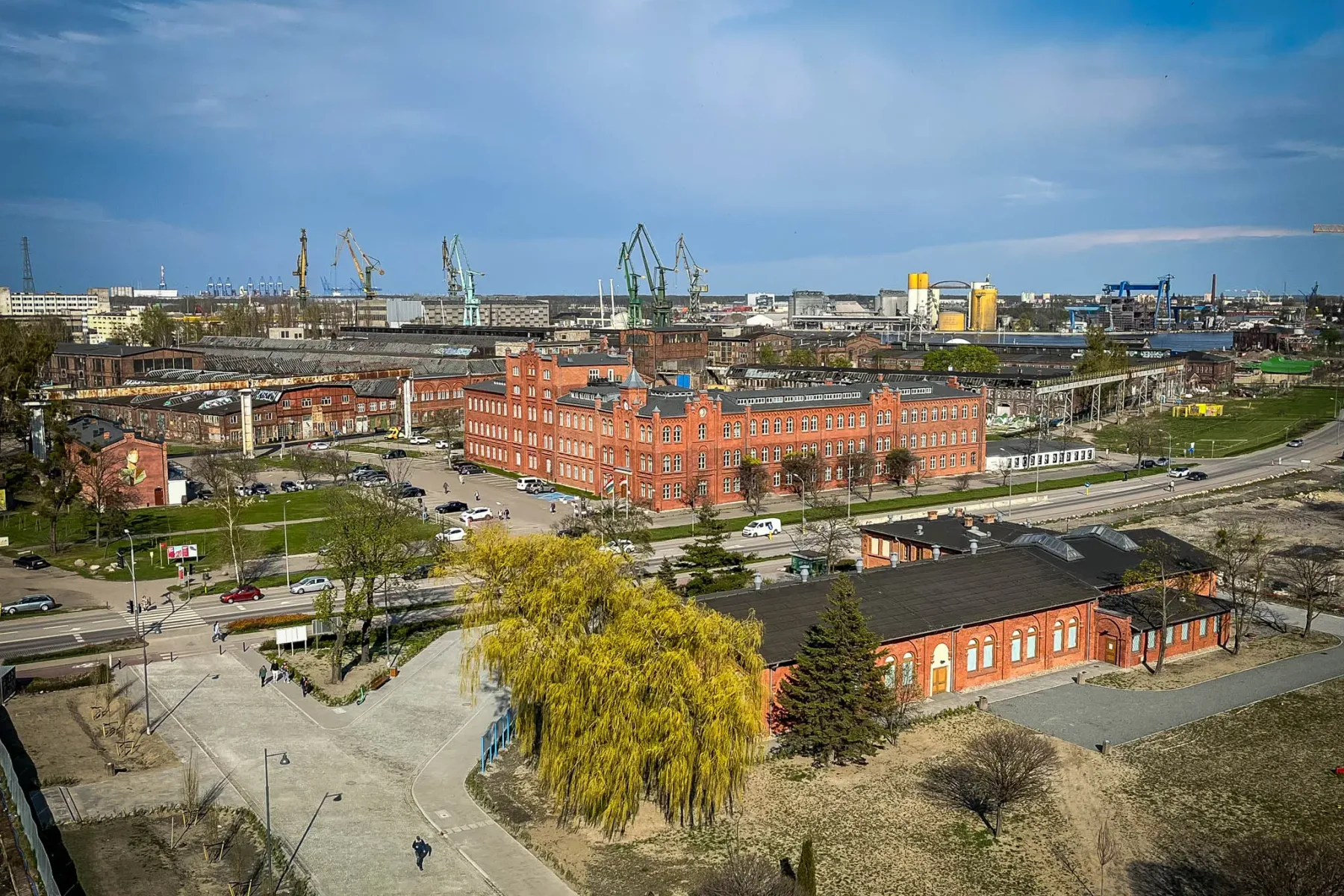 Aerial view of Gdańsk shipyard area with red brick buildings, roads, cars, and green industrial cranes under a blue sky.