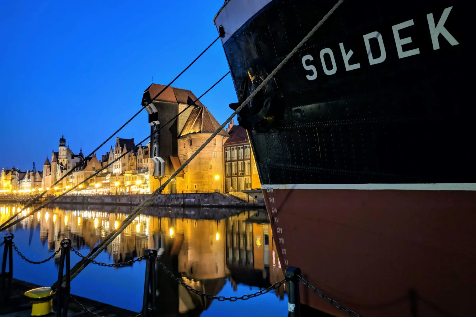 The black and red hull of MS Sołdek ship moored on a river, with illuminated historic buildings and the Żuraw crane reflec...