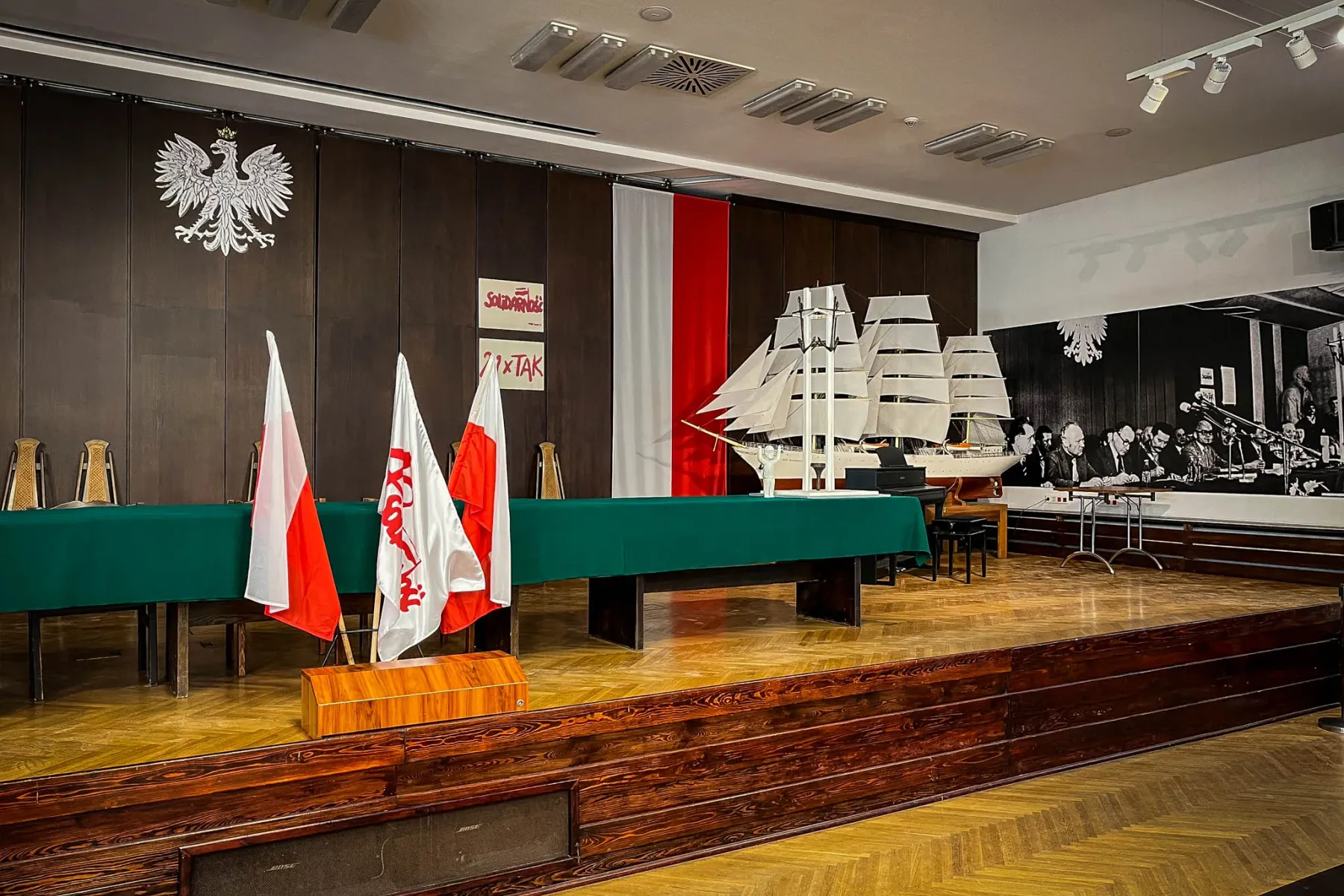 Conference stage with a green table, Polish flags, a model sailing ship, and a Polish eagle emblem on a dark wooden wall.