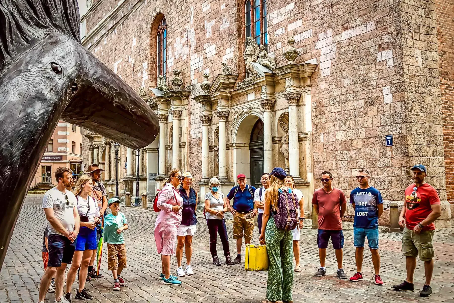Group of tourists listening to a guide on a cobblestone street in front of Riga's St. Peter's Church, with a sculpture on ...
