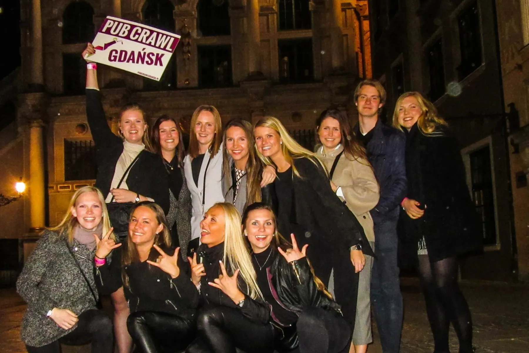 A group of young adults smiling and posing at night in front of a historic building. One person holds a sign reading 'PUB ...