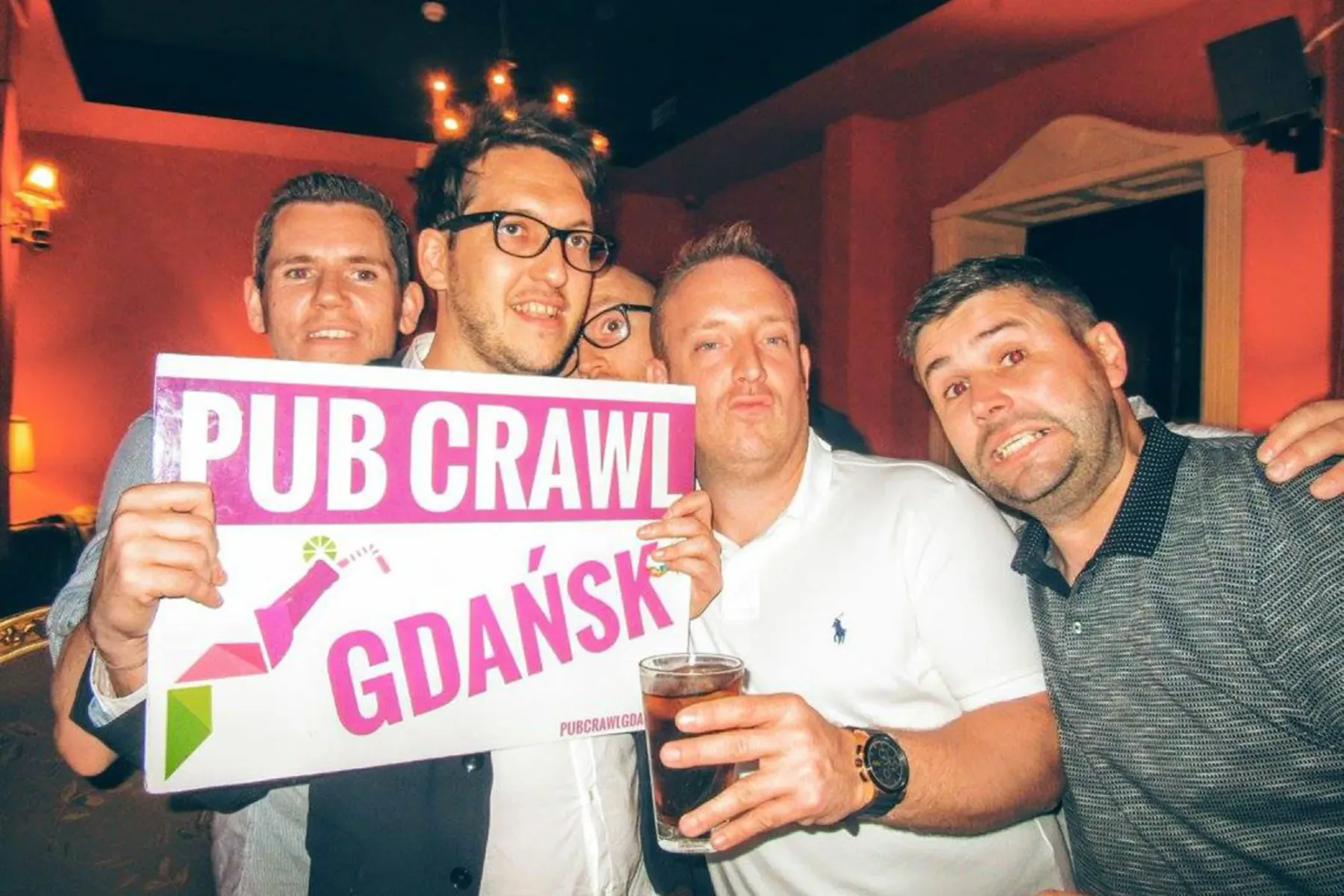 Four men in a dimly lit bar, three smiling, one holding a white sign that reads 'PUB CRAWL GDAŃSK' in pink letters.