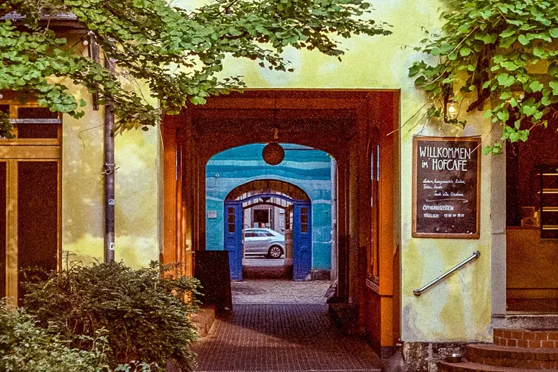 Paved courtyard leading to an archway, a building with yellow walls, windows, and a 'Willkommen im Hofcafé' sign. Green tr...