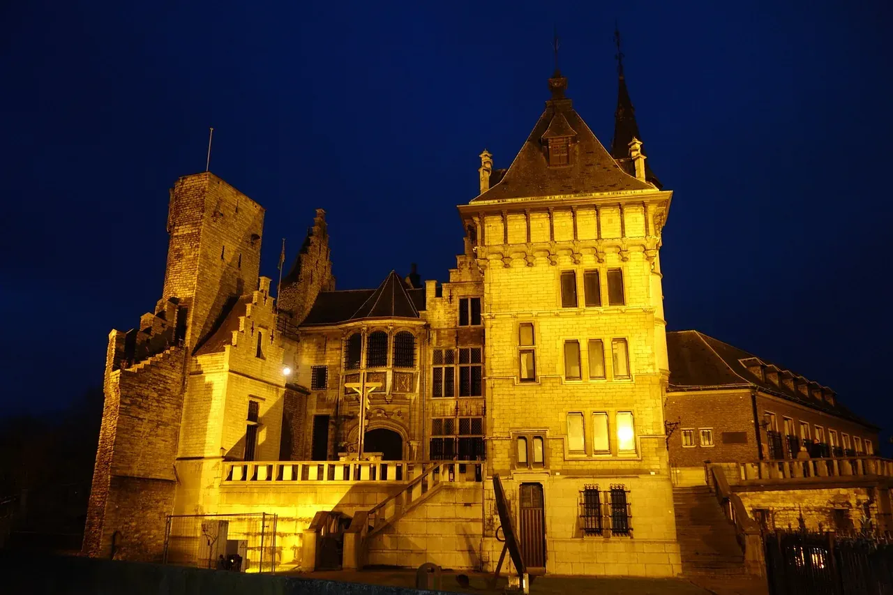 Gravensteen Castle in Ghent at night, illuminated with warm light, showcasing its stone walls, square tower, and gabled ro...