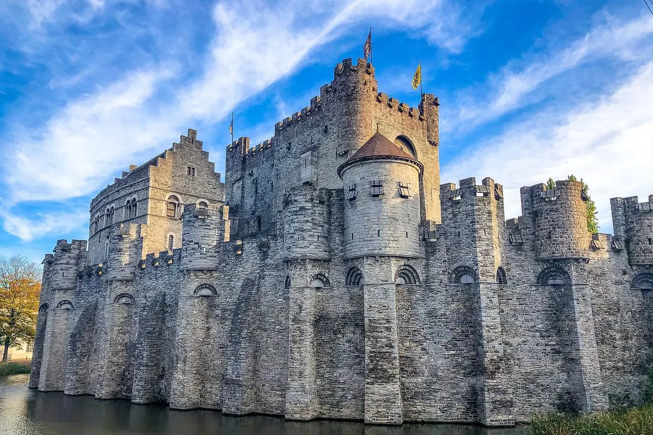 Stone medieval castle with multiple towers and battlements, surrounded by a dark moat under a blue sky with white clouds.