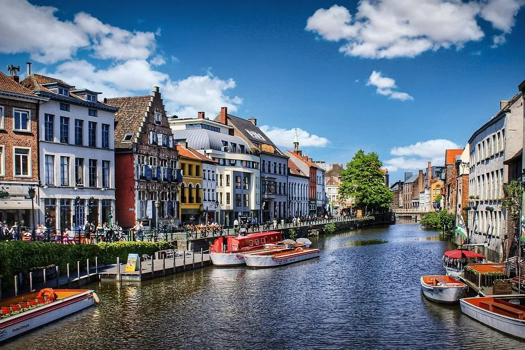 Historic buildings with gabled roofs line a sunny canal with several tour boats and people walking on the banks.