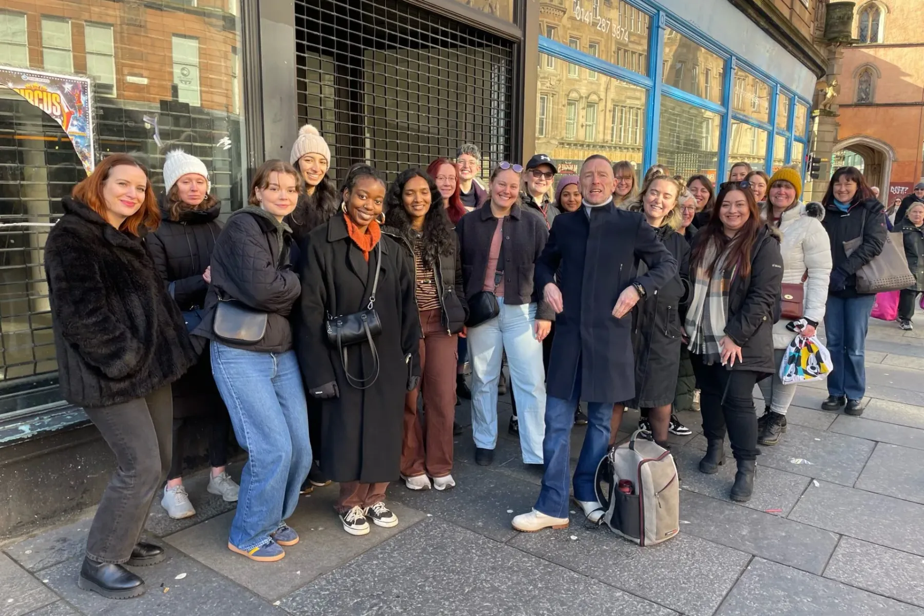 A diverse group of people and a guide smiling on a city street, standing in front of a shop with a metal security grate.