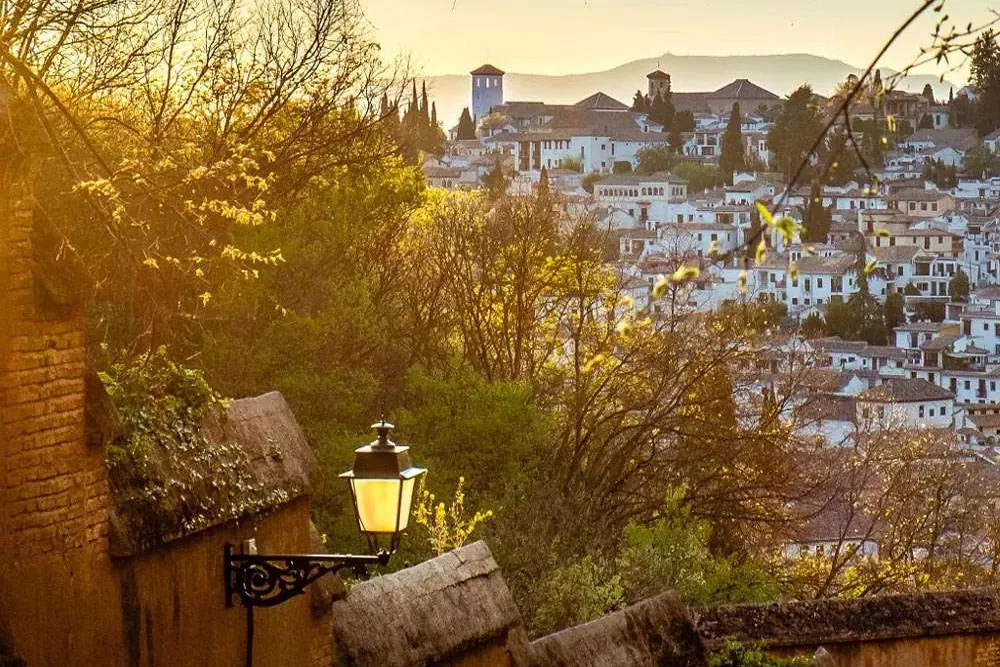Old brick wall with two lit street lamps, overlooking the white buildings and green trees of Granada's Albaicín at sunset.
