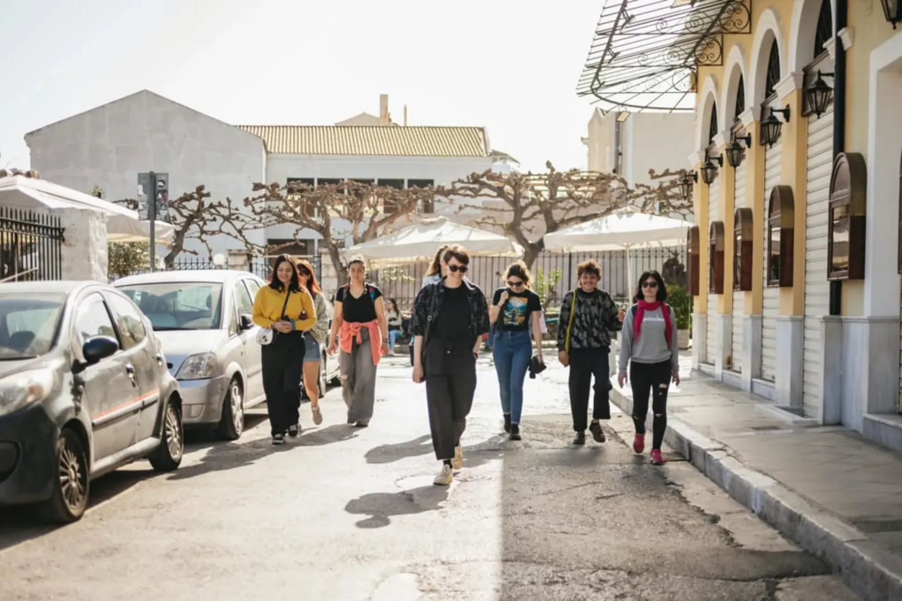A diverse group of tourists walking along a sunny street in a city, with parked cars on the left and arched buildings on t...