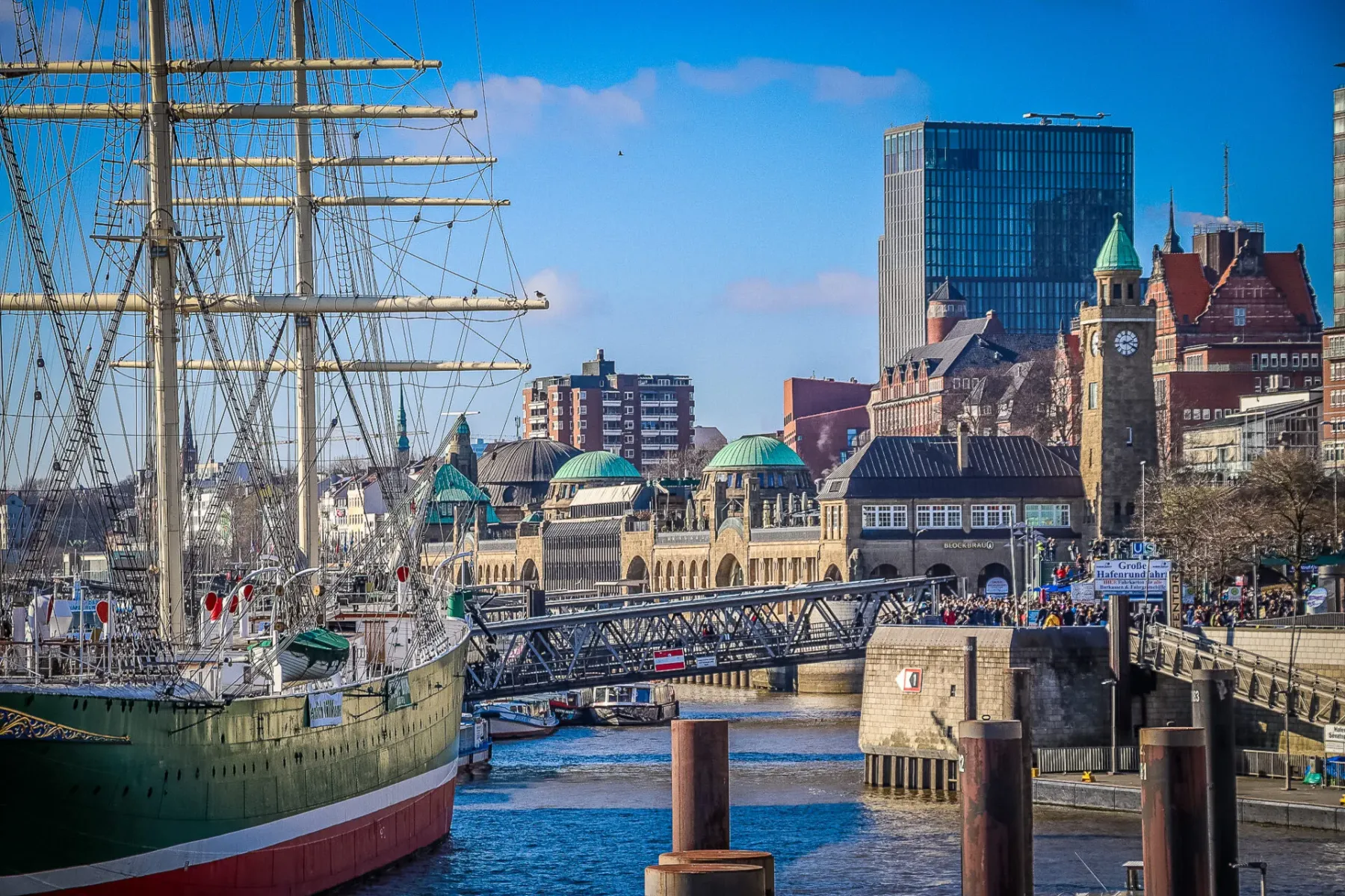 Large green and red tall ship docked in a harbor, with a bustling city waterfront and historic buildings in the background...