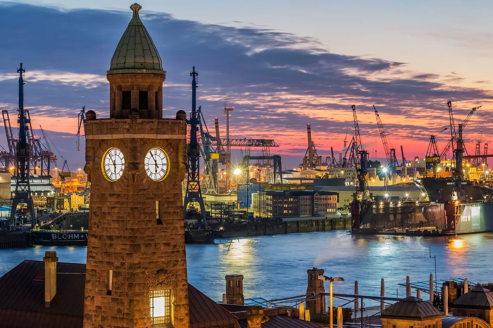 Stone clock tower with illuminated faces overlooking a bustling industrial harbor at dusk, with many cranes and ships in H...