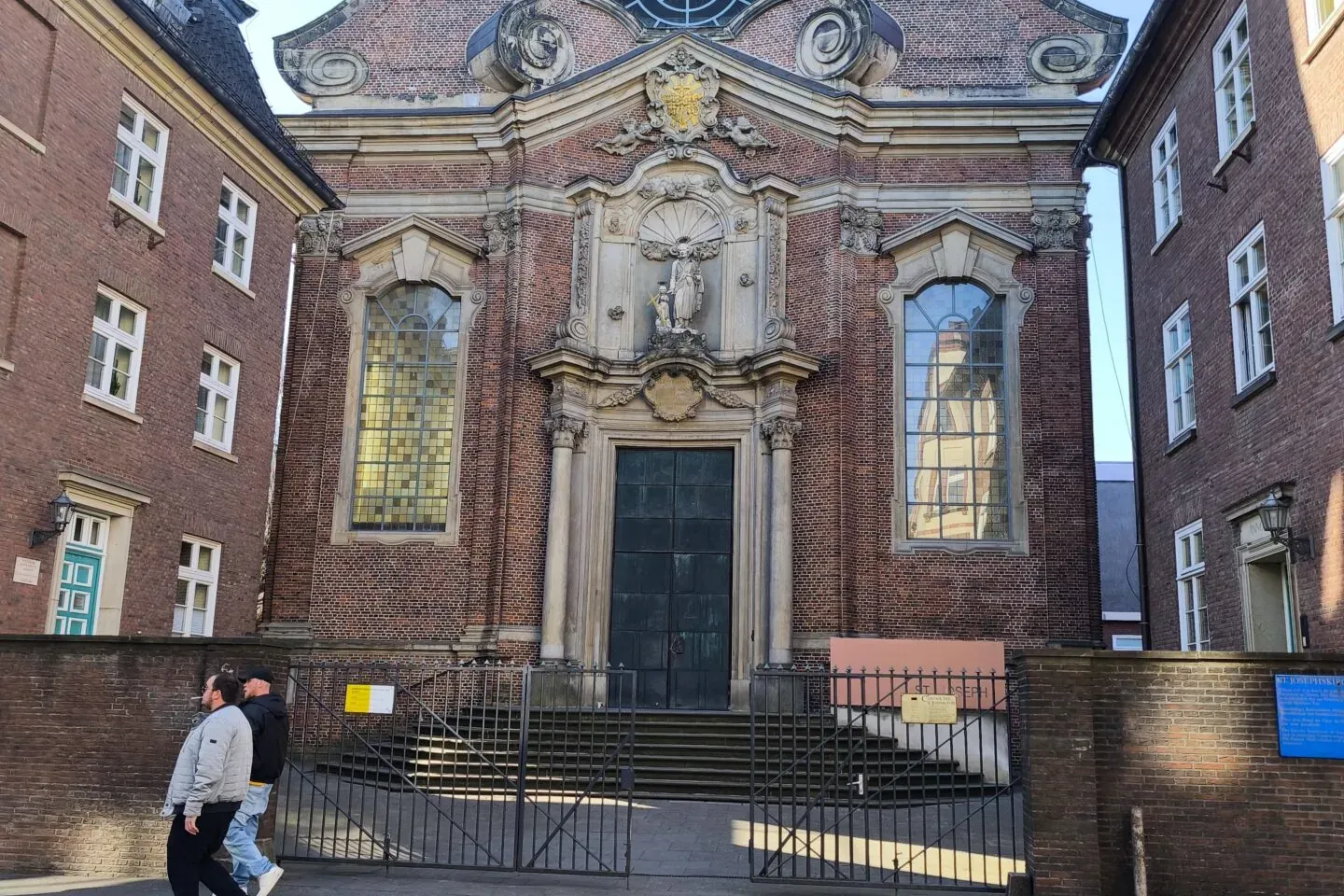 Baroque brick church facade with a large arched entrance, circular window, and cross, flanked by red brick buildings. Two ...