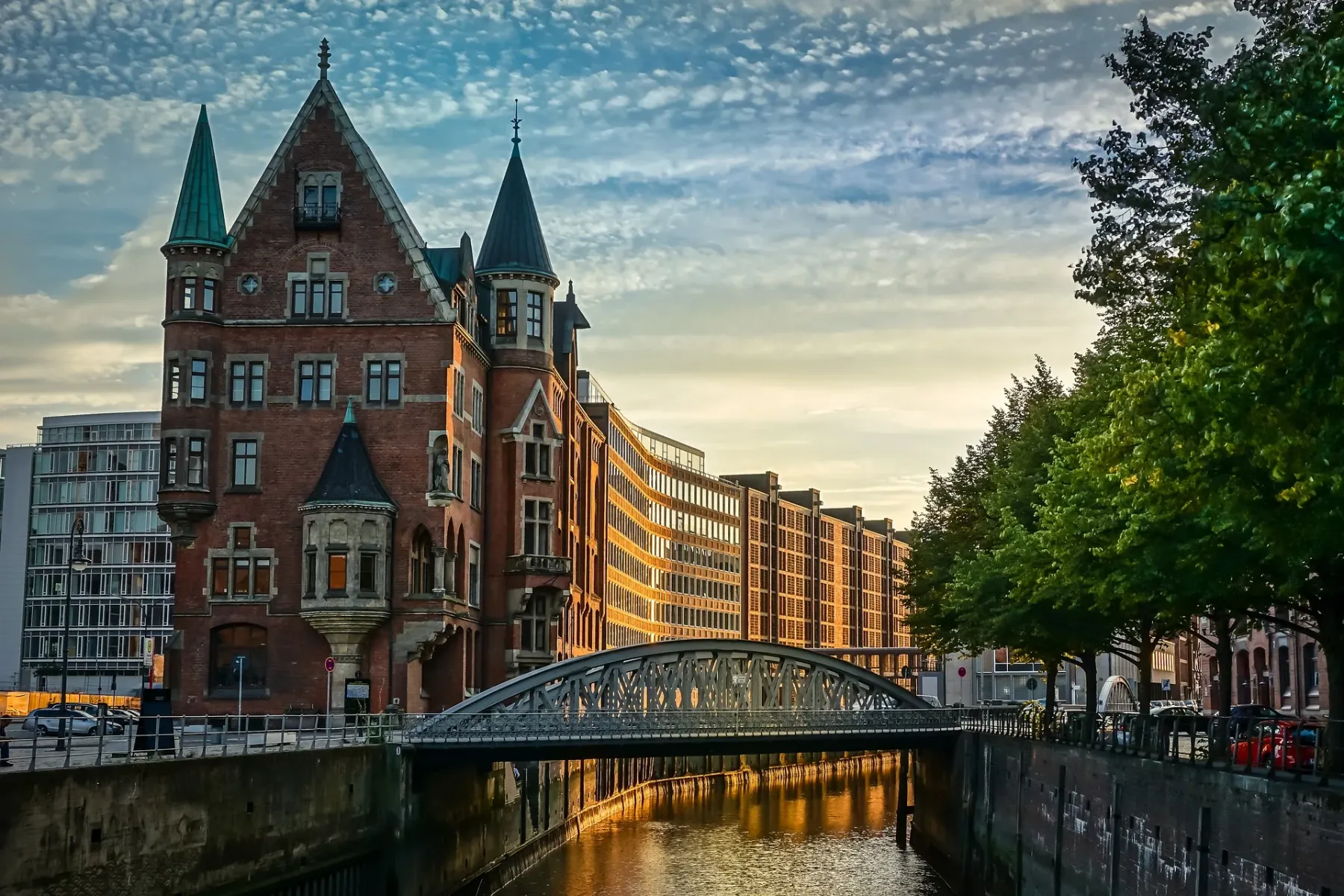 Historic brick building with turrets by a canal with an arched bridge. Reflections of buildings and a warm sky on the water.
