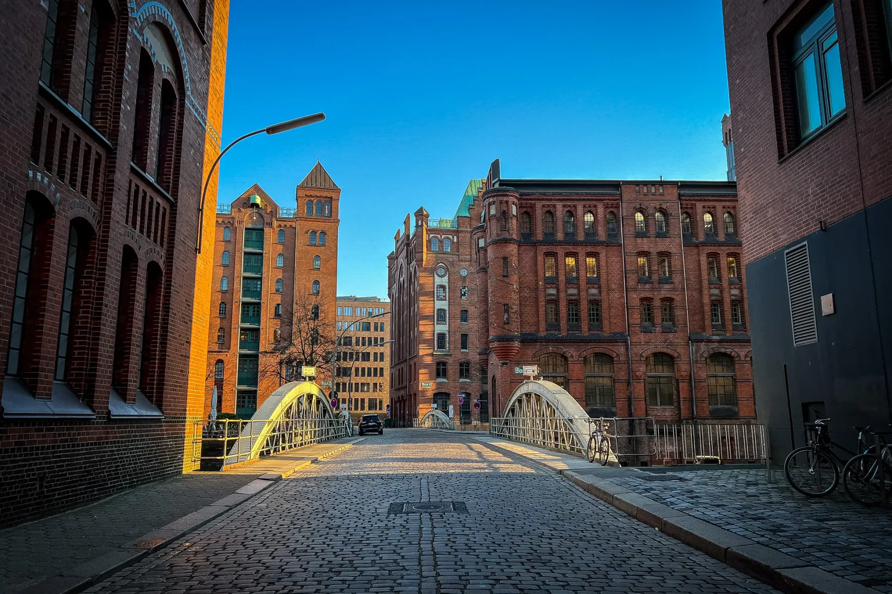 Cobblestone street leading to an arched white bridge, flanked by red brick warehouse buildings in Hamburg under a clear bl...