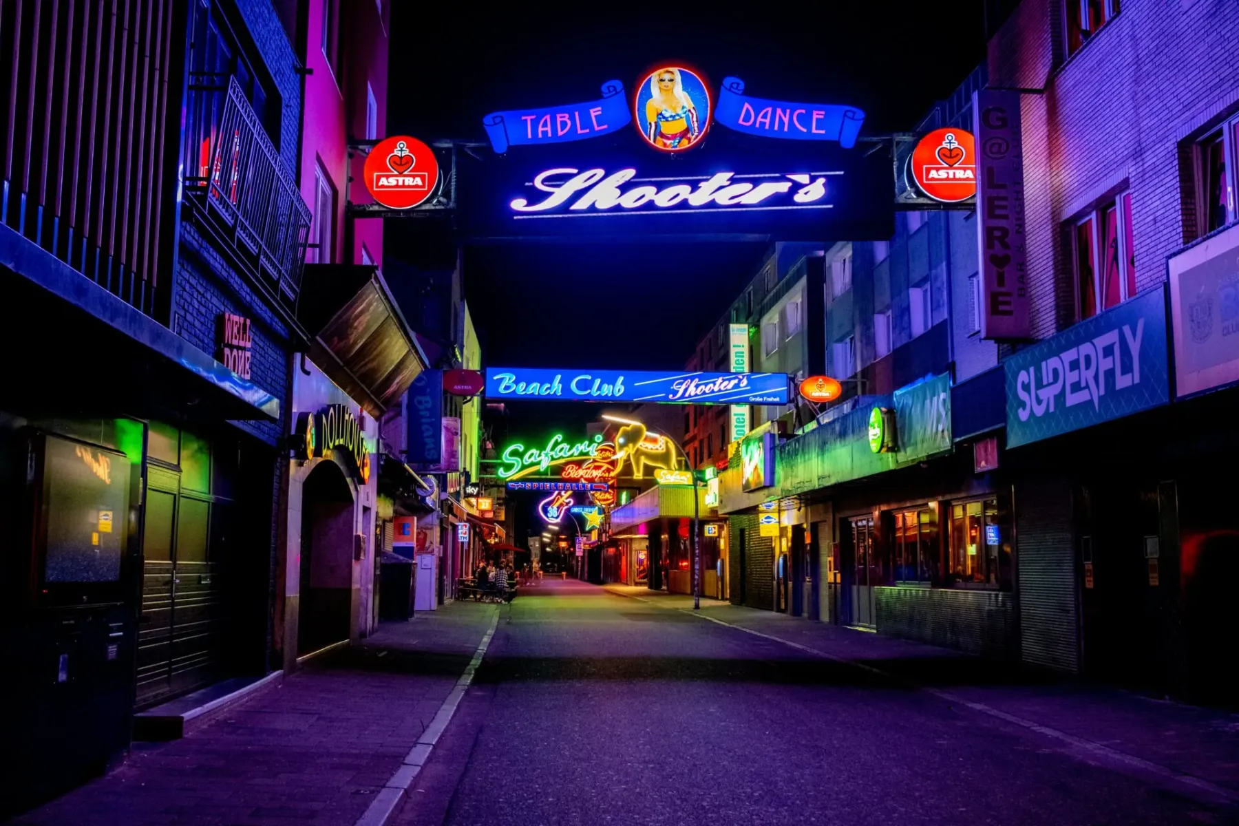 Neon-lit street at night in Hamburg, with various bar and club signs like "Shooter's" and "Safari" glowing purple and green.