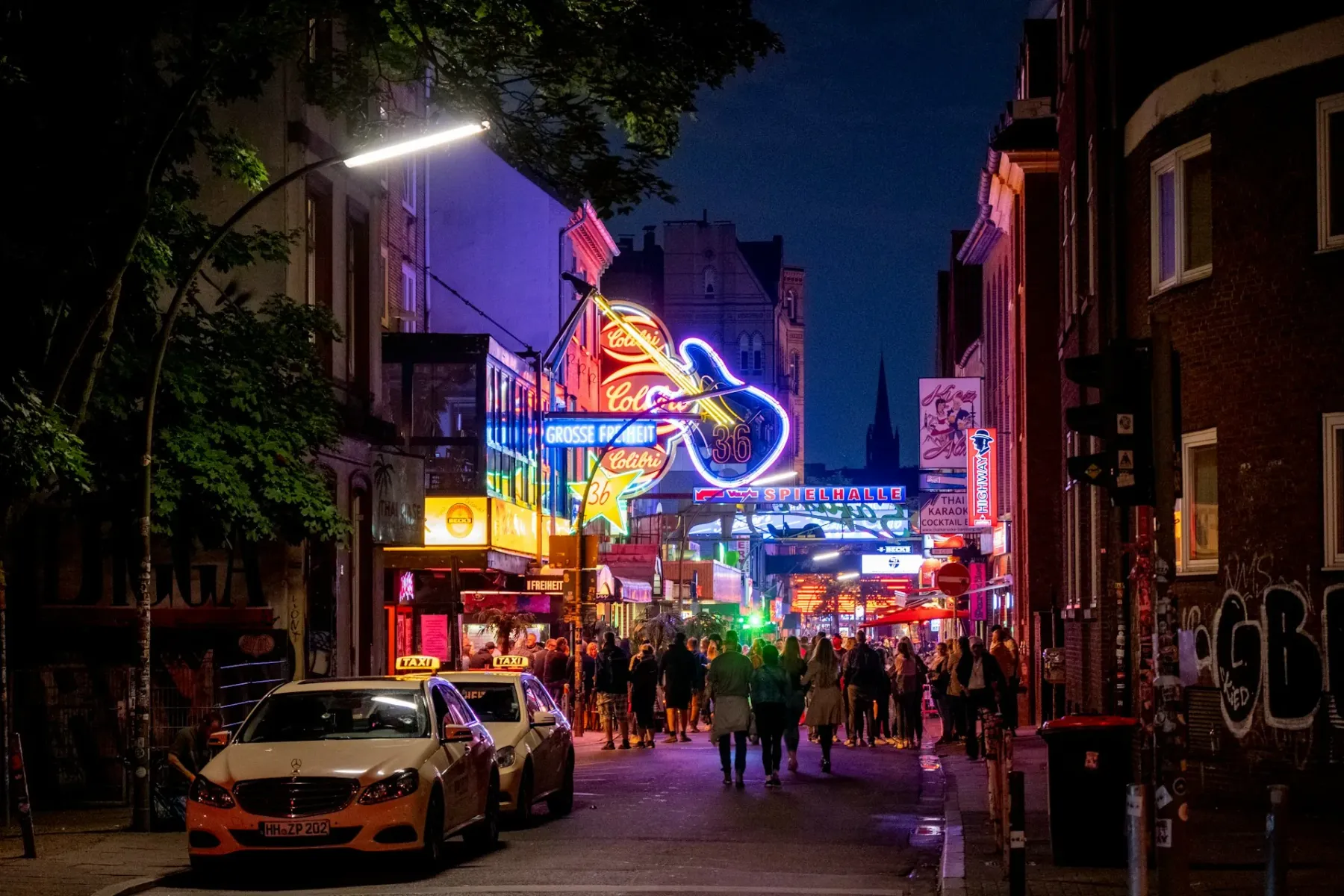 Night scene on Grosse Freiheit street in Hamburg with neon signs, people walking, and parked taxis. A church spire is visi...