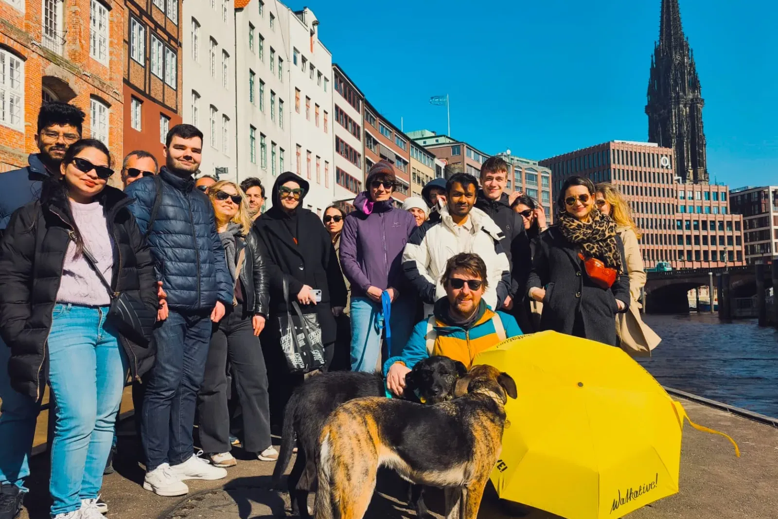 A diverse group of people on a walking tour next to a canal in Hamburg, with historic buildings and St. Nikolai Church spi...