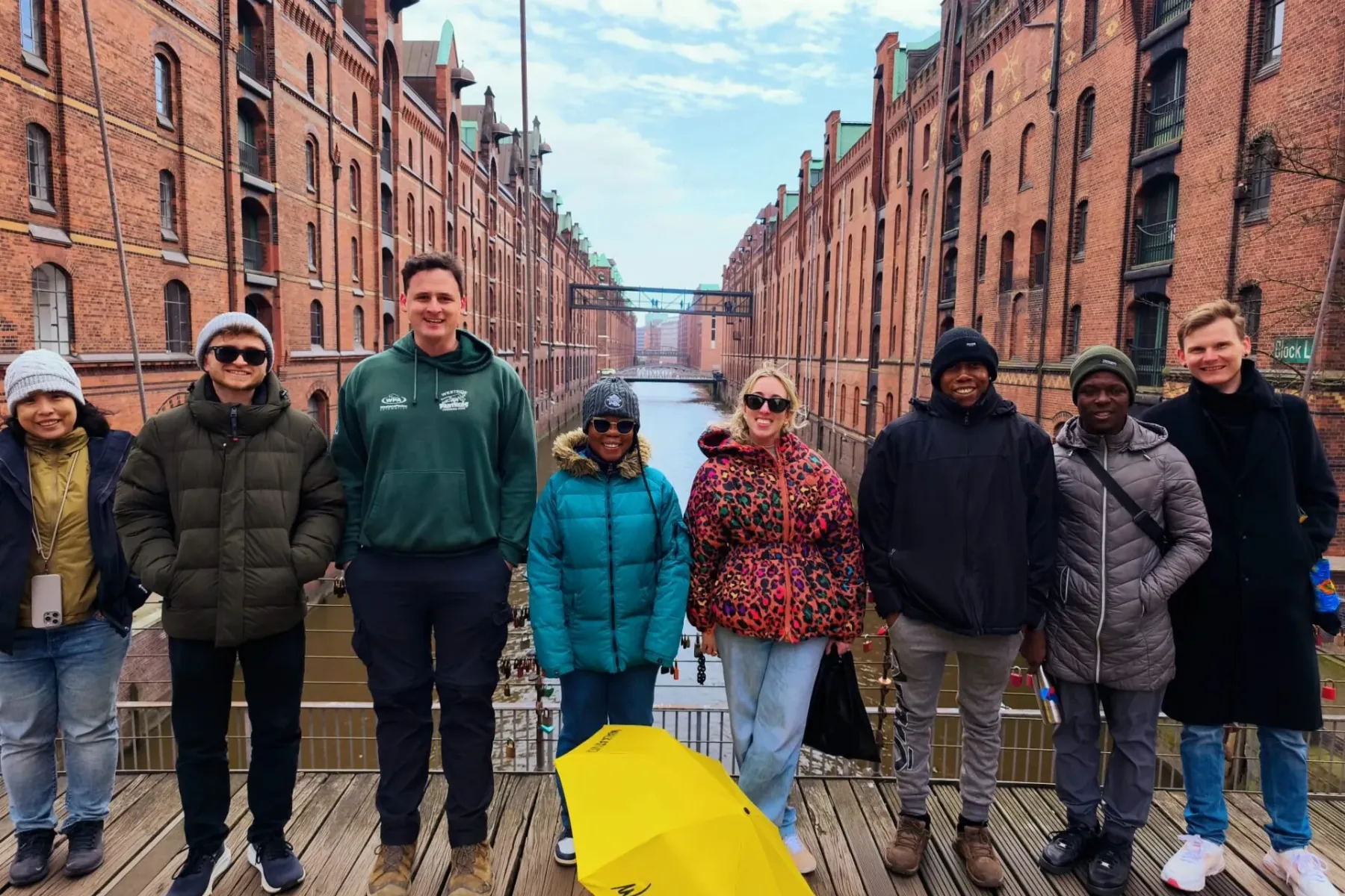 Group of diverse tourists and a guide standing on a wooden bridge in Hamburg's Speicherstadt, with red-brick warehouses an...