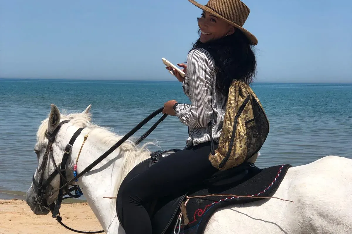Woman with a straw hat and backpack riding a white horse on a sandy beach next to calm blue sea under a clear sky.