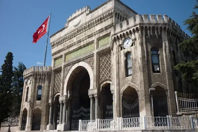 Ornate stone main gate of Istanbul University with a prominent Turkish flag flying, and a clock on a side tower.