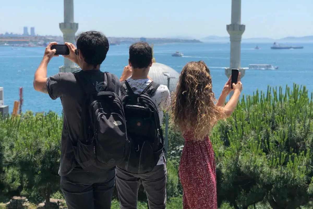 Three people photograph the Bosphorus strait, with minarets and a mosque dome in the background, under a clear blue sky.