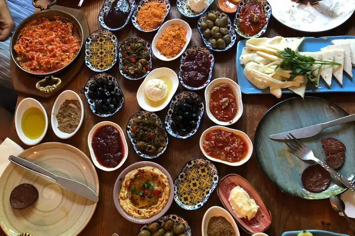 Overhead view of a large wooden table with a traditional Turkish breakfast spread: various small dishes of dips, olives, c...