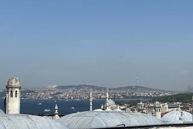 View over Istanbul's Bosphorus, showing city buildings, distant mosques on hills, and boats on the water under a clear sky.