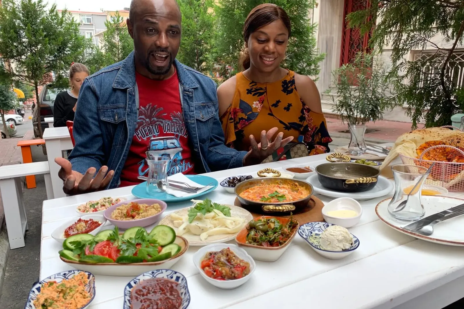 A man and a woman at an outdoor table overflowing with a variety of Turkish breakfast dishes like eggs, meze, and fresh ve...