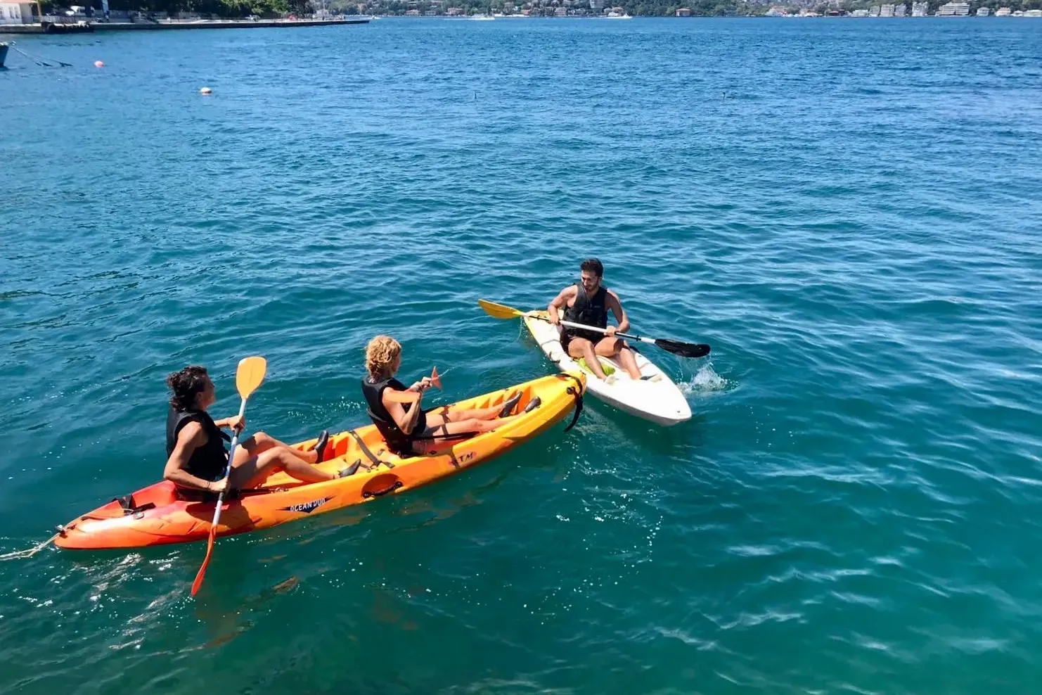 Three people in life vests on two kayaks on blue water. Two are in an orange double kayak, one in a white single. Green tr...